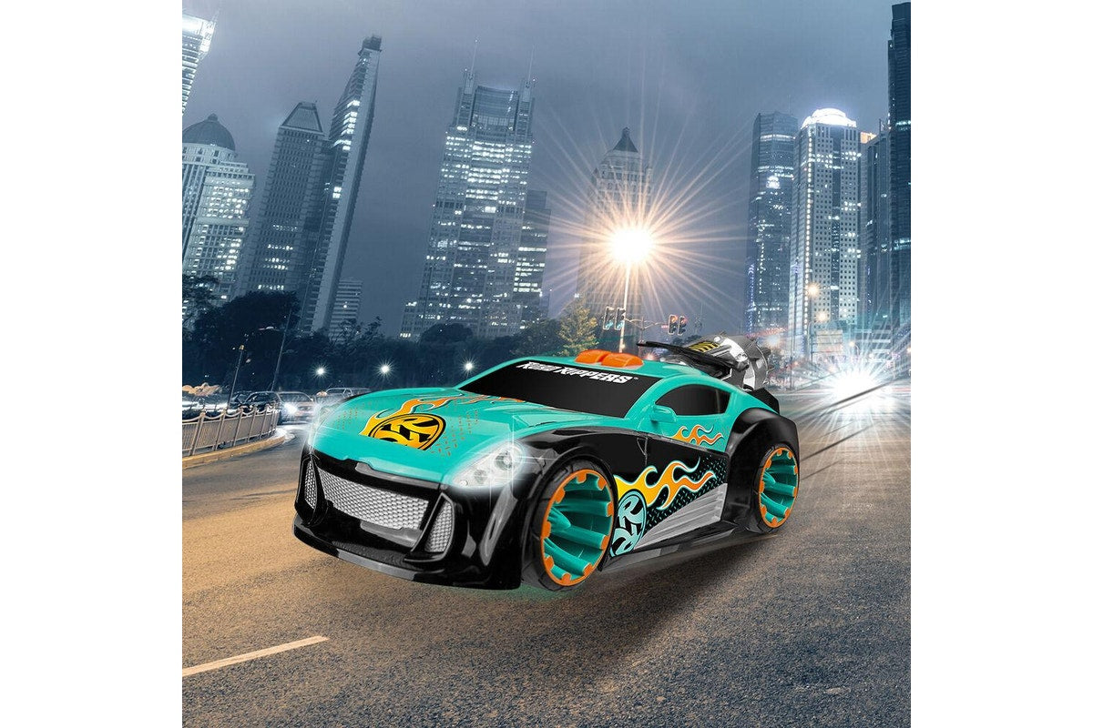 A teal and black toy sports car with orange and yellow flame decals, shown driving on a city street at night with illuminated skyscrapers in the background.