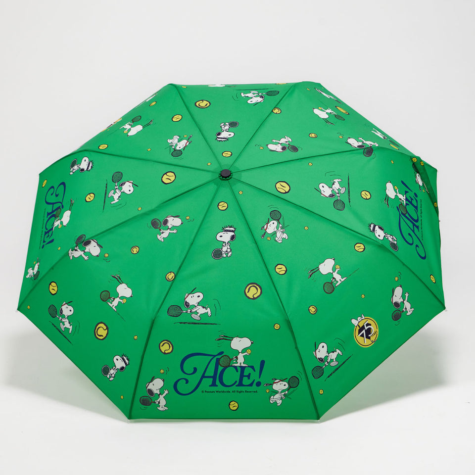 A green folding umbrella with a repeating cartoon print of Snoopy and his dog friends, tennis rackets, tennis balls, and the word 'Ace!' in blue script near the bottom.