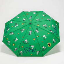 A green folding umbrella with a repeating cartoon print of Snoopy and his dog friends, tennis rackets, tennis balls, and the word 'Ace!' in blue script near the bottom.