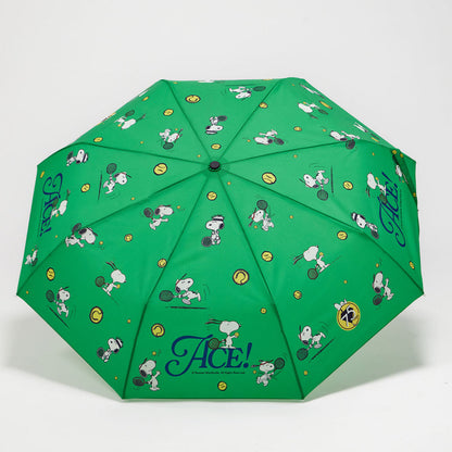 A green folding umbrella with a repeating cartoon print of Snoopy and his dog friends, tennis rackets, tennis balls, and the word 'Ace!' in blue script near the bottom.