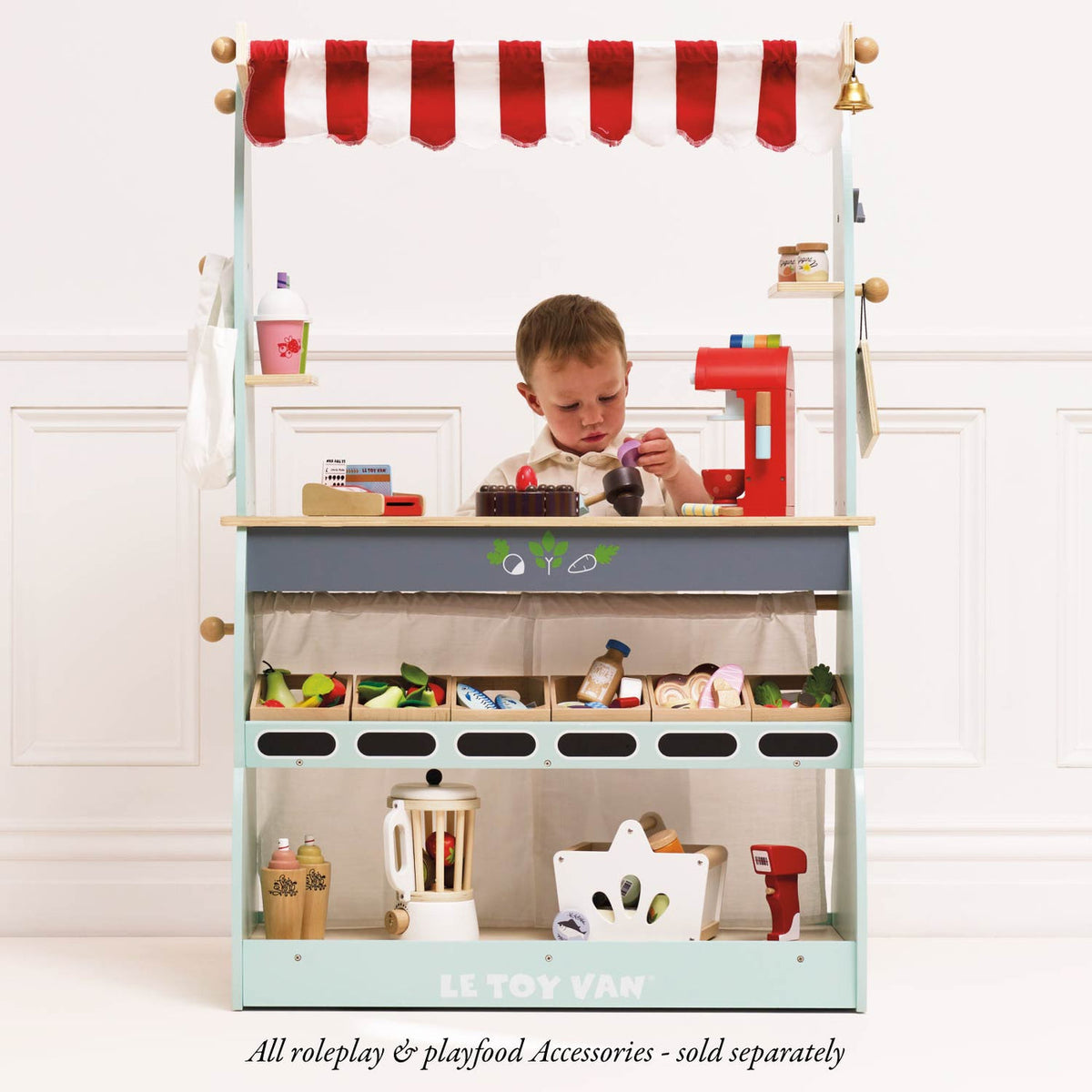 Child enjoying imaginative café role play behind the Le Toy Van Wooden Shop & Café Market Stand, featuring a striped canopy, display shelves, pretend groceries and toy coffee machine. Play accessories sold separately.