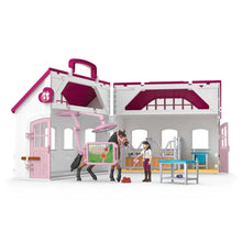 A pink and white horse stable toy set with an open stable, a horse in a stall, a small figure of a woman, and grooming accessories, photographed on a clean white background.