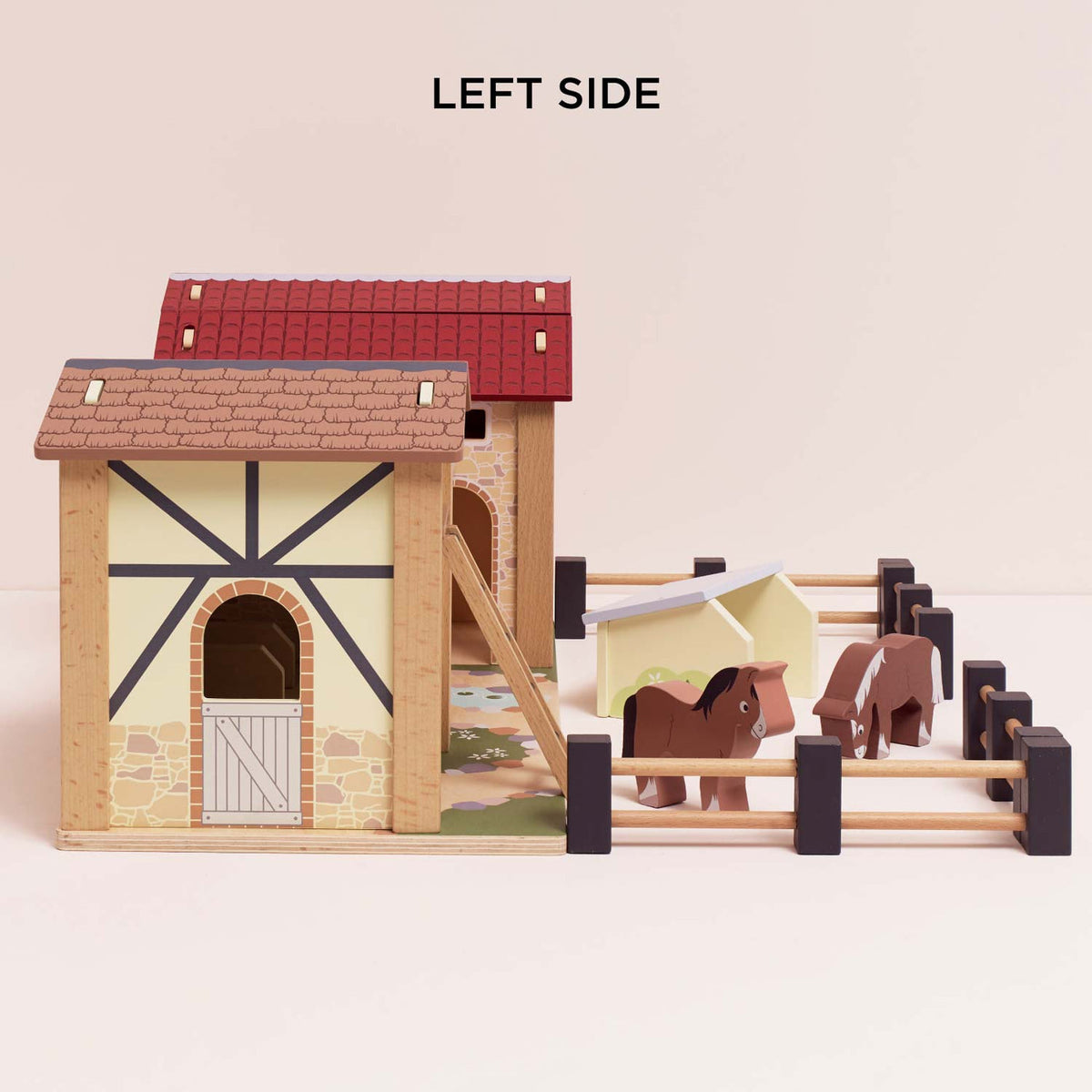 Left side view of Le Toy Van Wooden Farmyard Set featuring the timber-style barn with stable door, tiled roof, wooden ladder, pigsty and two horses inside a fenced paddock.