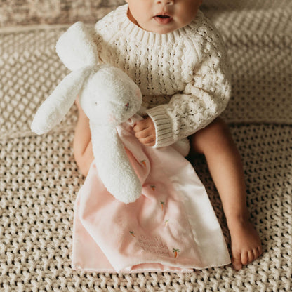 Baby wearing a cream knitted jumper holding a soft white bunny comforter with a pale pink blanket and embroidered carrots.