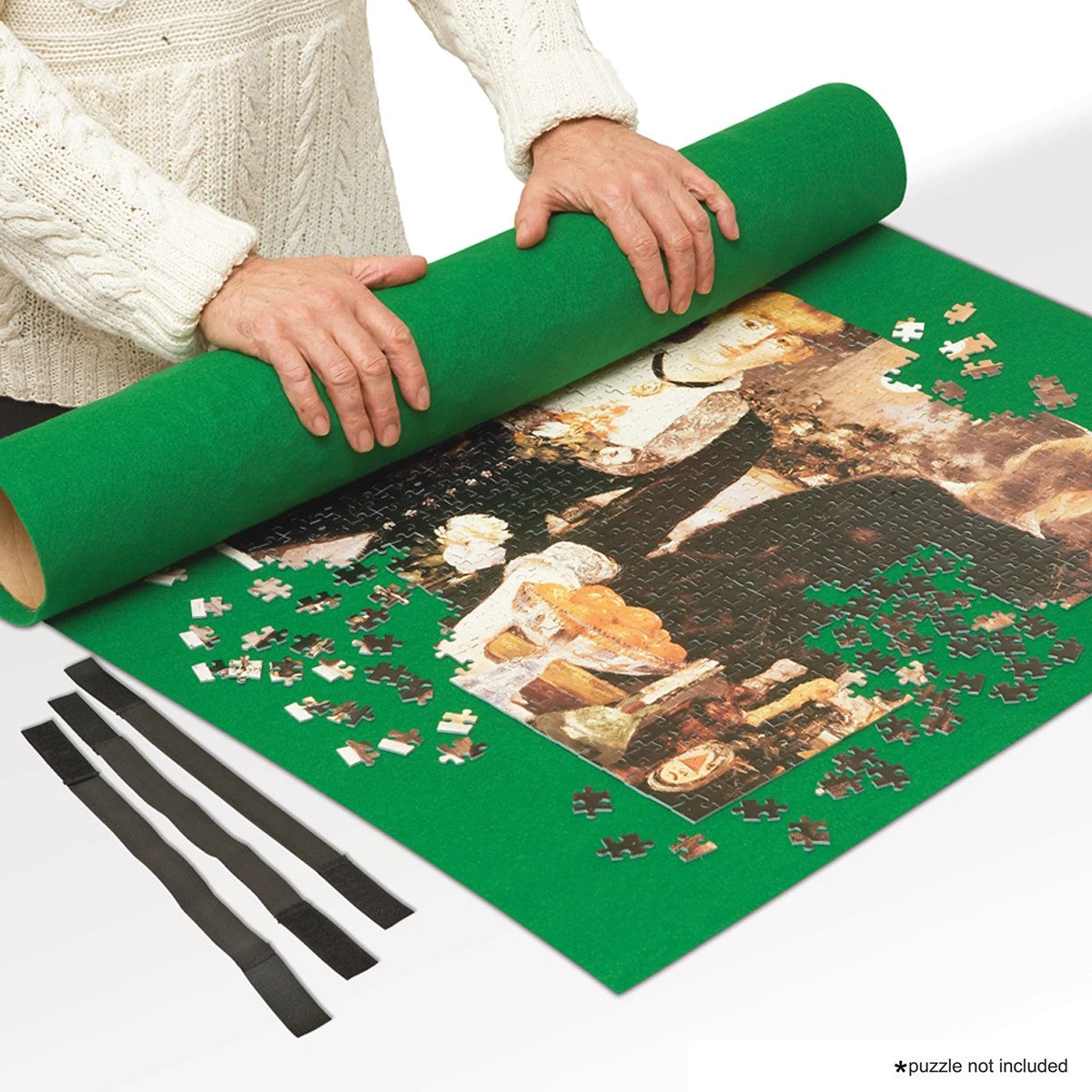 A person in a cream cable-knit sweater is unrolling a green felt puzzle mat over a partially assembled jigsaw puzzle. Three black elastic bands are laid out beside the mat on a white background.