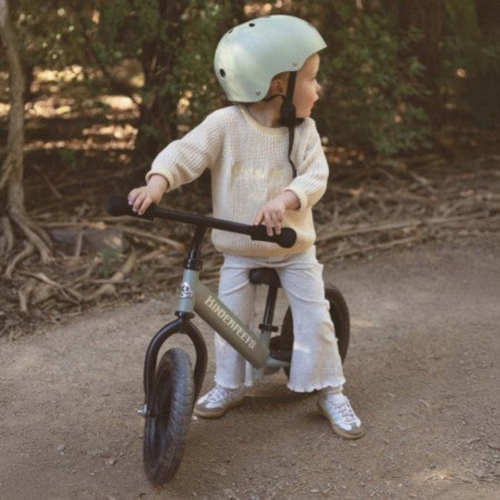 Child wearing a helmet seated on the Kinderfeets Trail Balance Bike in Silver Sage outdoors