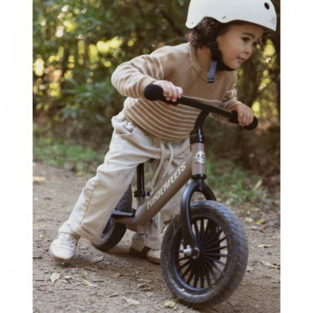 Young child wearing a white helmet riding a beige Kinderfeets balance bike with black wheels and handlebars along an outdoor dirt path.