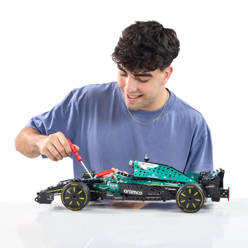 A man in a blue t-shirt is assembling a green and black Technic Formula 1 toy car with a red screwdriver, set against a clean white background.
