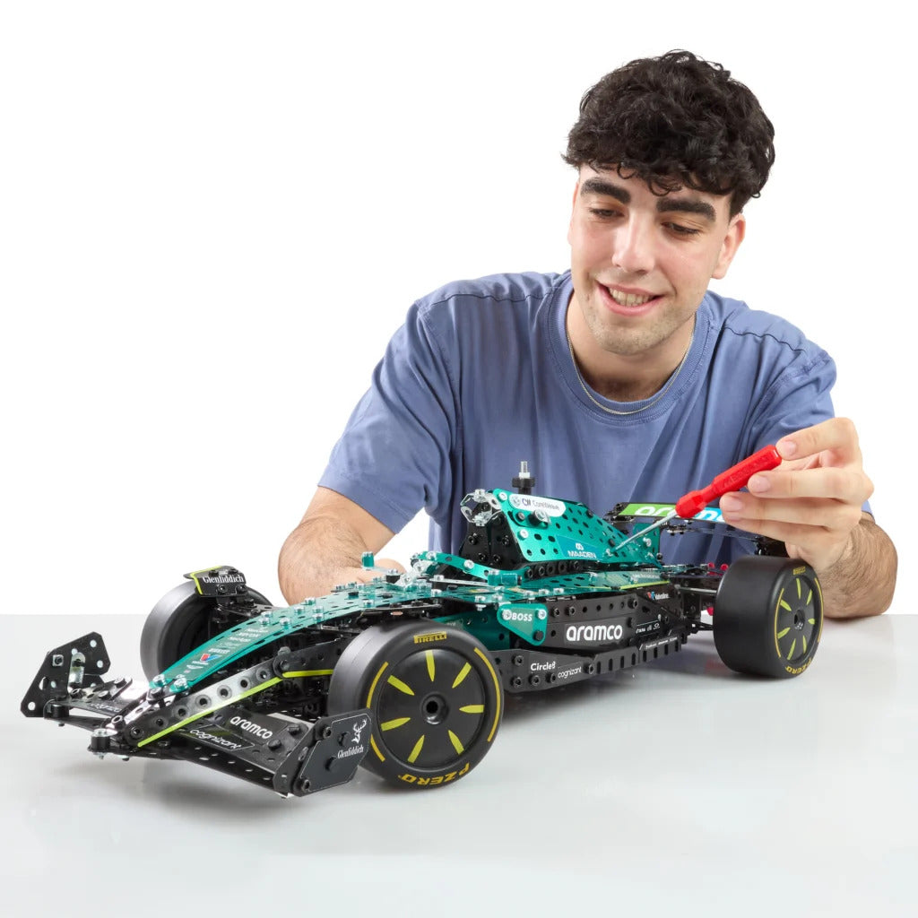 A young man smiling and holding a red tool next to a green and black LEGO Formula 1 car on a light gray surface against a white background.