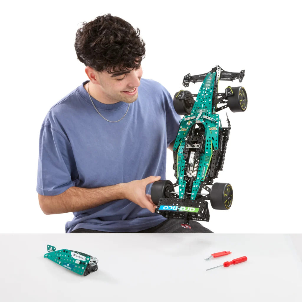 A man in a blue t-shirt holds a green and black robotic race car chassis built from building blocks, with a small loose electronic module and two red-handled screwdrivers on a white table in front of him against a white background.