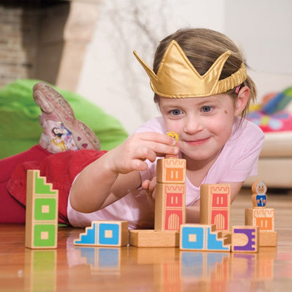 Young girl wearing a gold princess crown playing SmartGames Camelot Jr, placing the princess figure onto a colourful wooden castle puzzle with stairway blocks, towers and a knight figure.