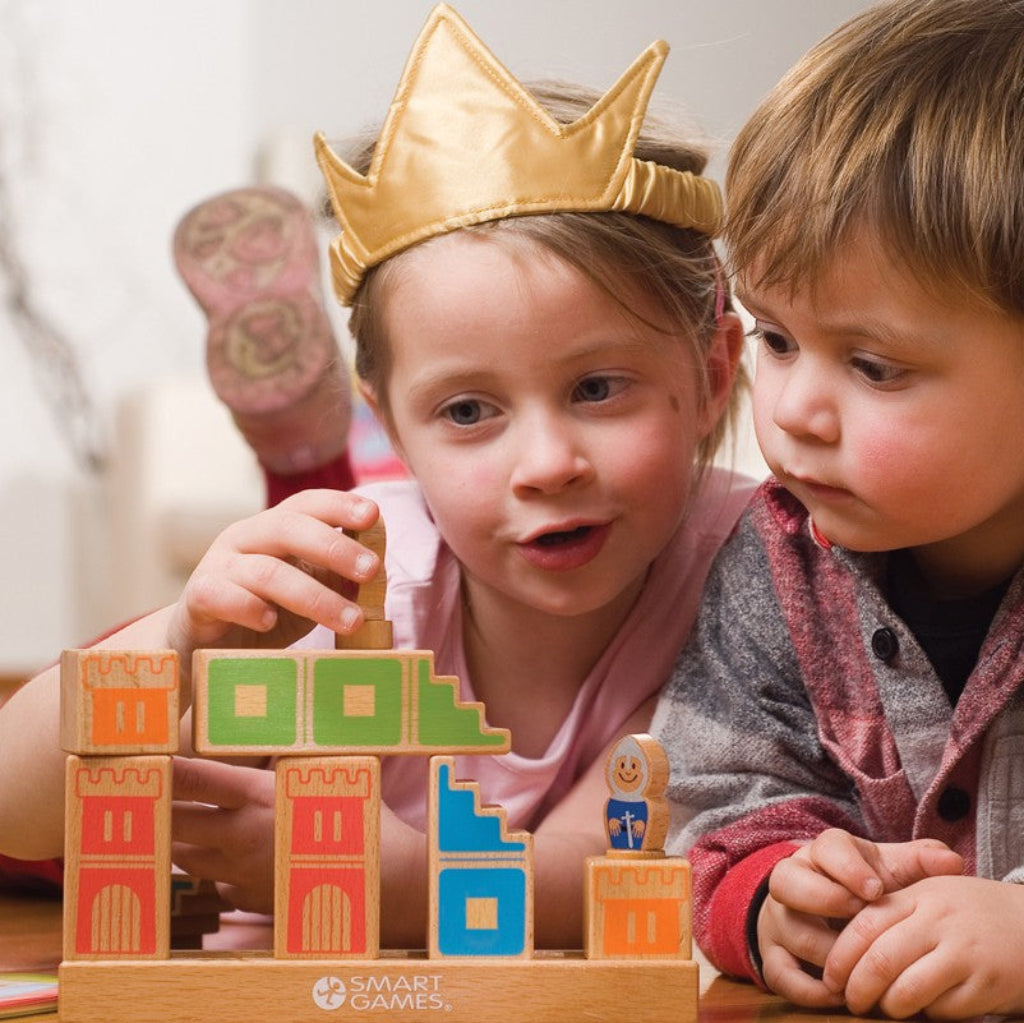 Two children playing SmartGames Camelot Jr wooden logic puzzle, arranging colourful castle blocks and stairways to create a path for the knight, while one child wears a gold princess crown.