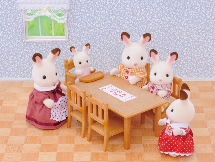 Toy figurines of rabbits around a table in a room with wallpaper and window.
