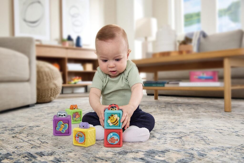Baby sitting on a play mat knocking over a tower of stacked VTech Baby Pop-a-Blocks activity blocks